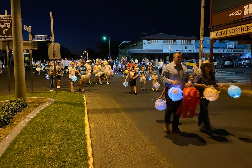 A group of people walk down a street at nighttime holding paper lanterns.