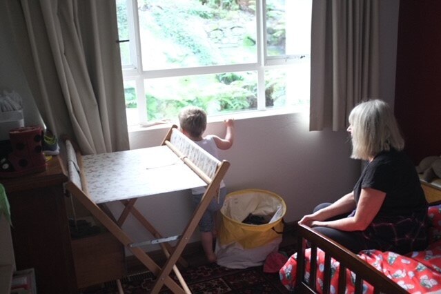 Part of Kate Flood's nursery, her baby is looking out the window and her mother is sitting on the bed and watching.