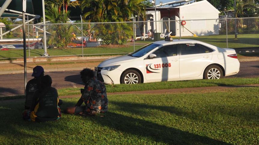 Three Aboriginal people sit in the shade next to a tropical taxi rank.