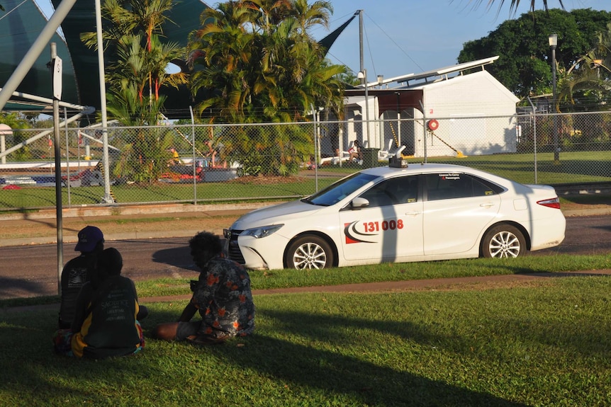 Three Aboriginal people sit in the shade next to a tropical taxi rank.