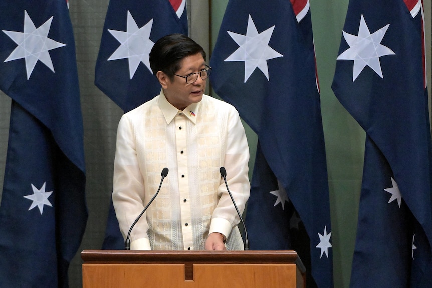 A man stands at a lectern in front of Australian flags.