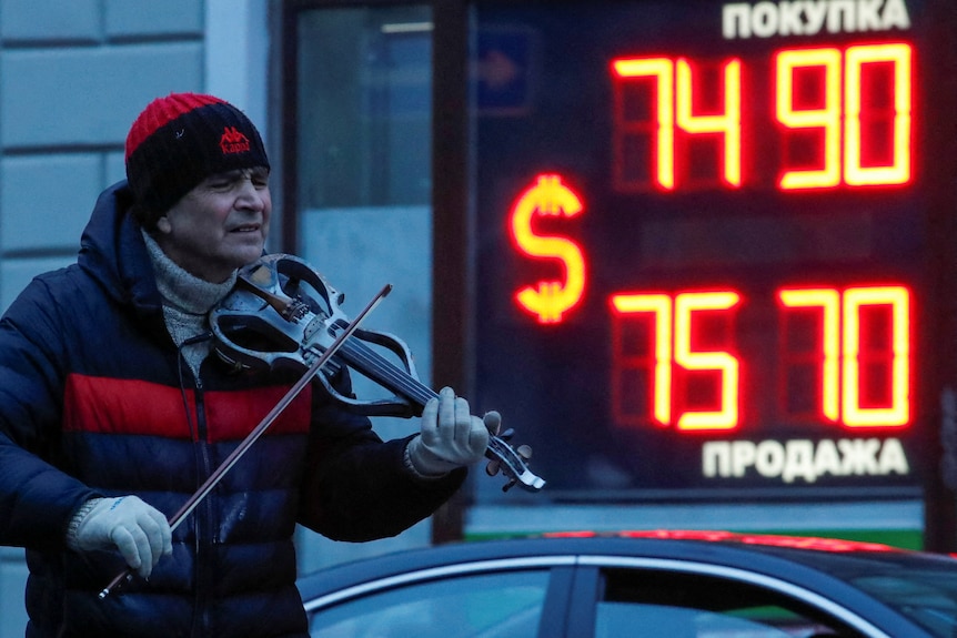 A man in a beanie, gloves and coat plays the violin while standing next to a board with LED numbers