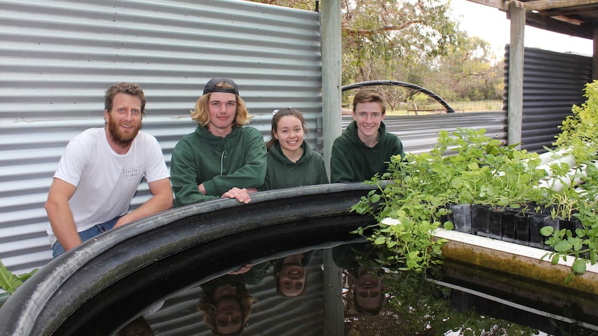 A male teacher and teenage students squat next to a water tank containing pots with green plants.