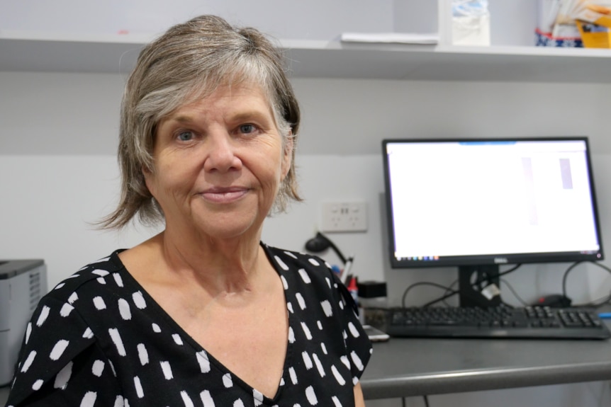 a woman in a black and white blouse with a computer behind her sits in a consult room, looking at the camera