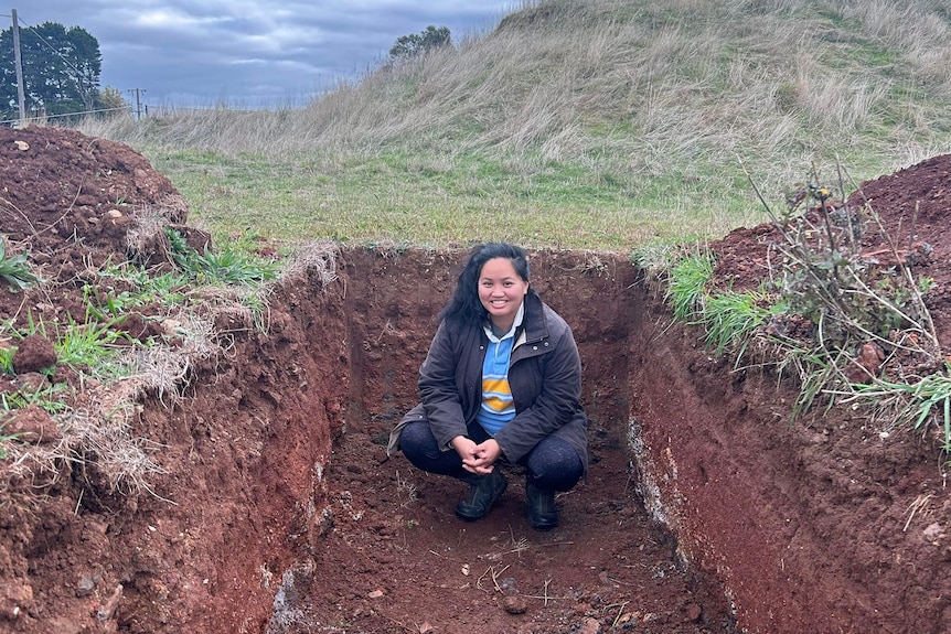 A Filipino-Australian woman, Kim, squats in a soil pit like a shallow grave cut out of grassy land.