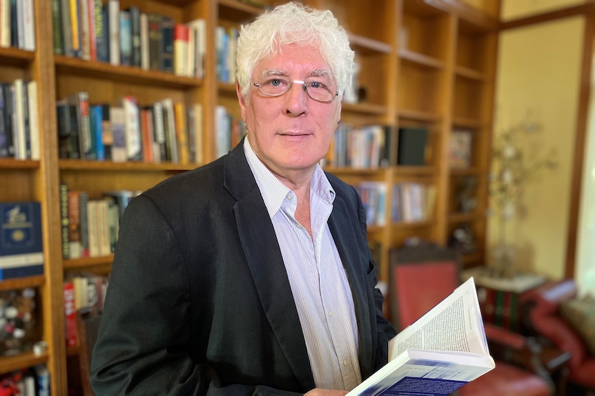 Professor Charles Sampford smiles at the camera while holding a book and standing in an office.