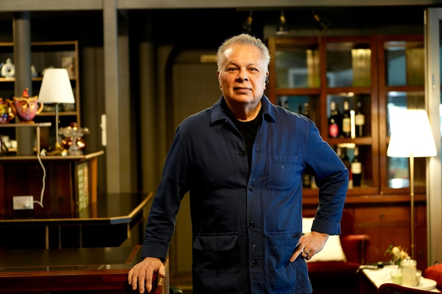 A man with light brown skin wearing a blue overshirt and black tshirt standing in a restaurant with his hand on his hip