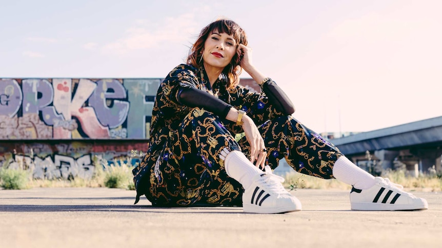 AROHA sits on concrete in front of graffiti. She wears a black tracksuit with gold detail and white sneakers with white socks.