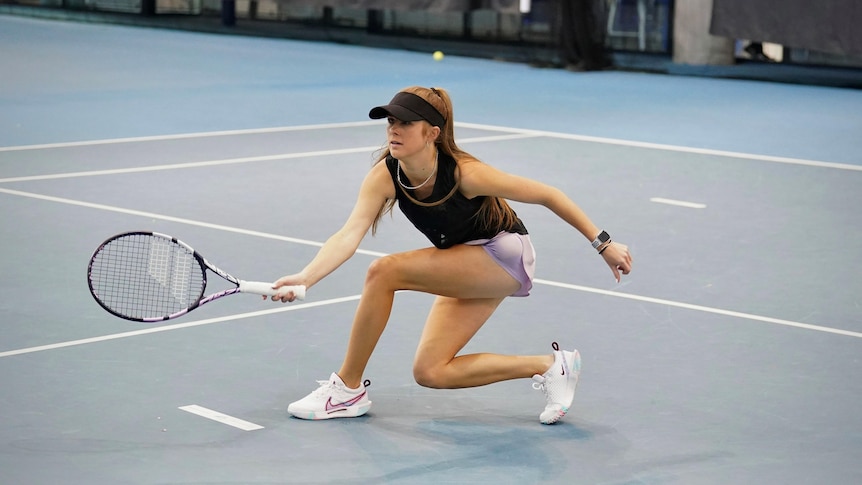 A young female tennis player, wearing a brown visor, lavender shorts,  brown top, plays on a tennis court..