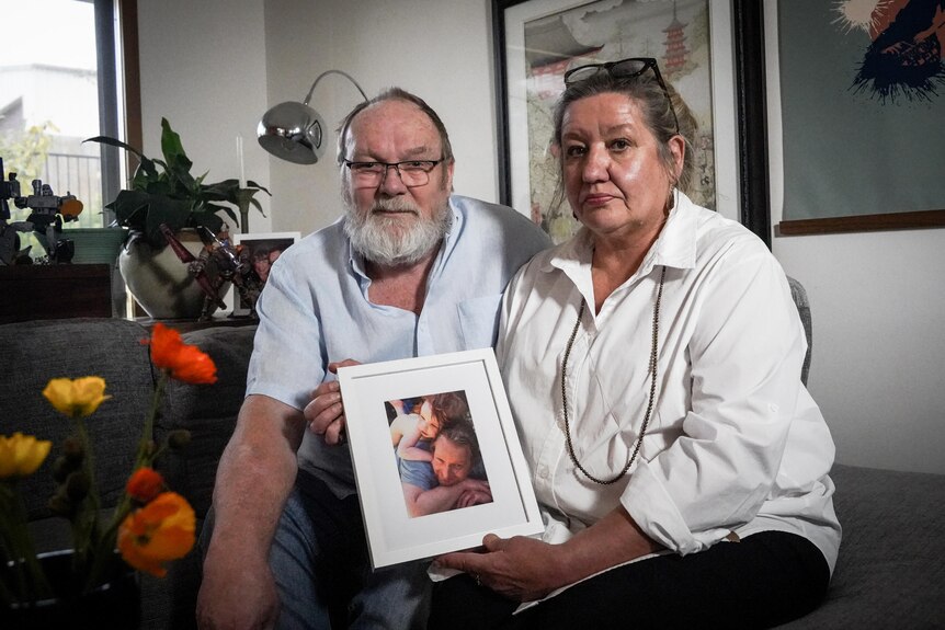 A man and a woman sit on a couch holding a photo of a man and his daughter, they look directly into the camera