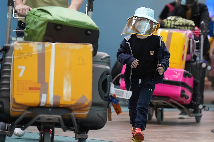 A child wearing a face mask and shield walks next to a luggage trolley inside an airport