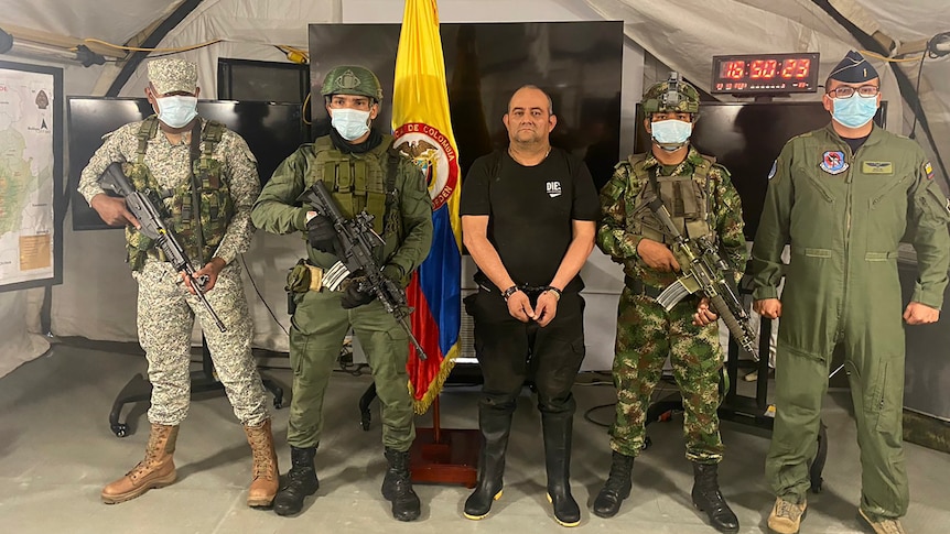 A man in handcuffs stands between four soldiers with guns. 