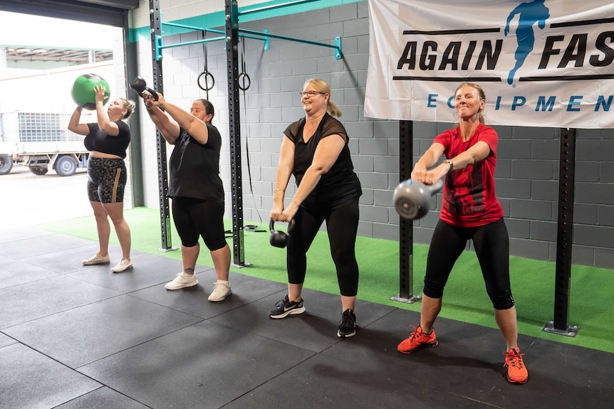 Four women stand in a line doing kettlebell swings and lifting medicine balls.