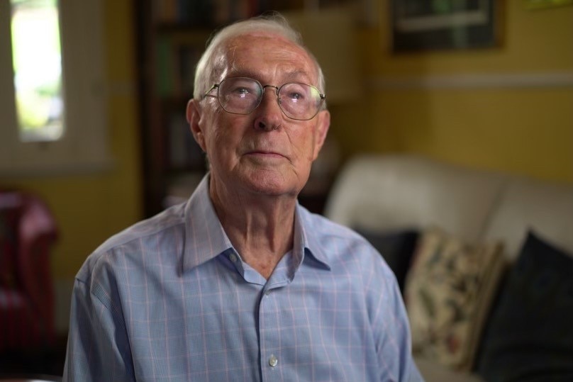 A man with grey hair and glasses inside a house looks into the camera. 