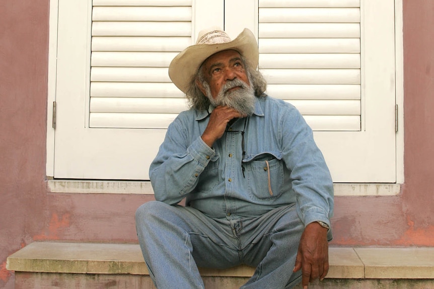 Bob Randall sits on a doorstep and looks up wearing a broad brimmed hat and denim shirt
