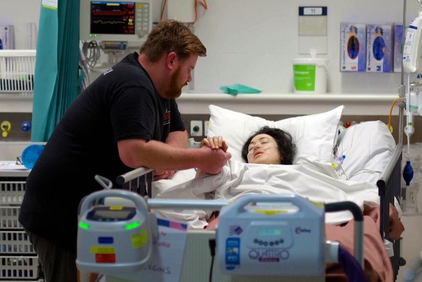 A man standing next to a woman who's in her hospital bed, holding her hand