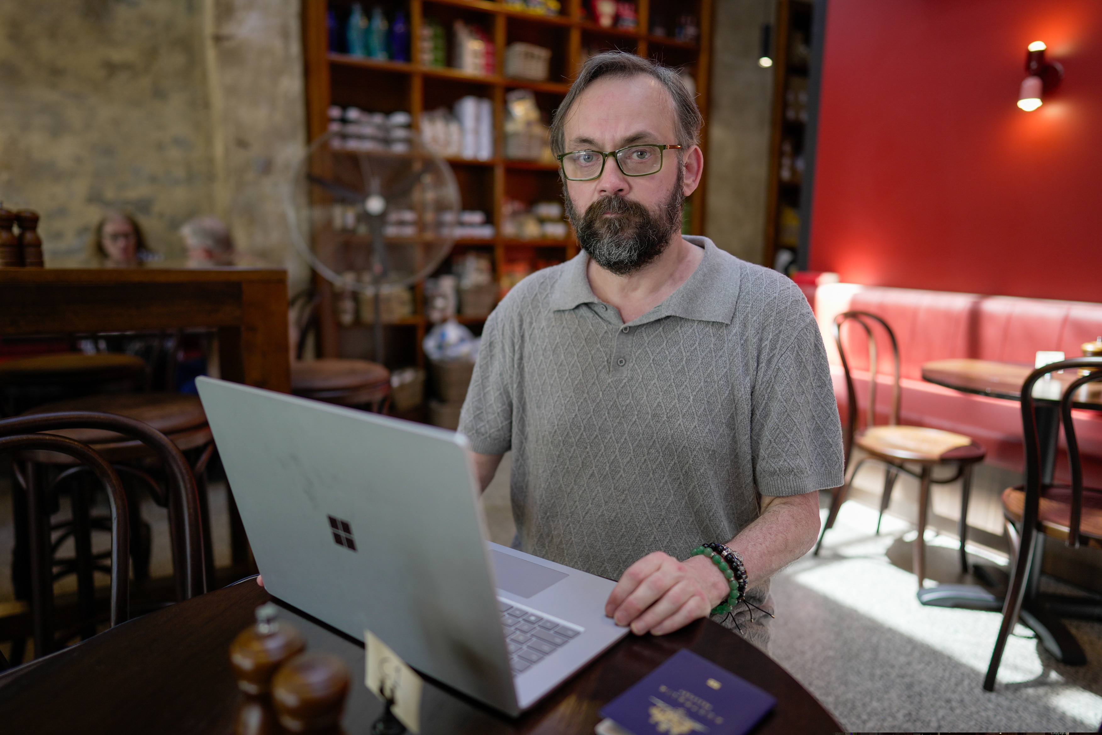 A man in a grey shirt and beard looks at a laptop in a cafe.