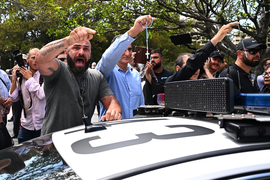 A man shouting, near a police car, next to another man holding a string of beads