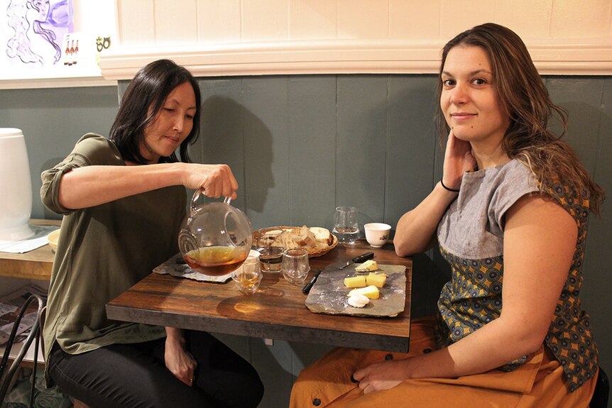 Two women sitting at a table in a cafe, pouring tea into cups beside a cheese board and basket of bread.