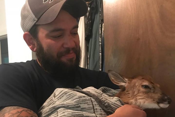 A tattooed man with a beard and trucker's cap cradles a fawn in a blanket