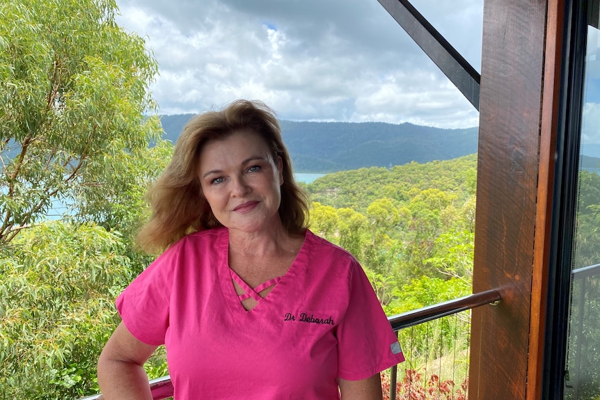 A woman stands in a pink doctors scrubs with the ocean and trees behind her