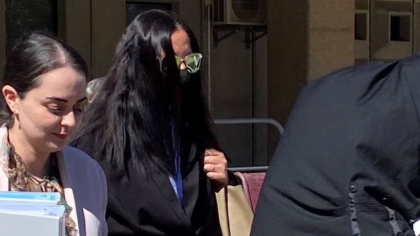 A woman walks between two lawyers. She has long black hair that covers her face. She wears sunglasses and a face mask.