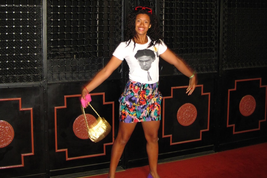 A teenage girl stands in front of a black wall while wearing a white shirt, multicoloured skirt and gold bag.