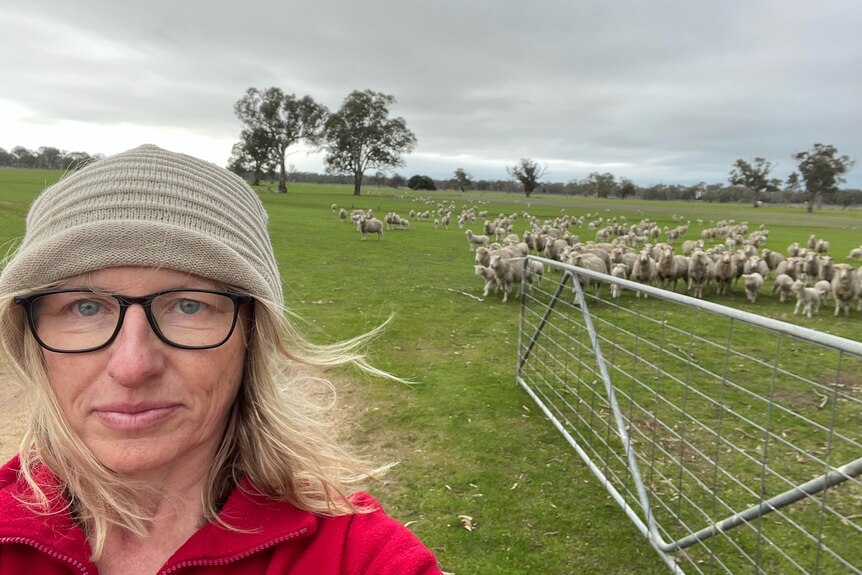 A woman wearing a beanie and glasses poses in a sheep paddock for a selfie.