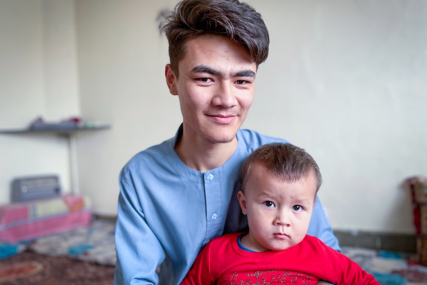 A teen boy grins while holding a toddler on his lap 