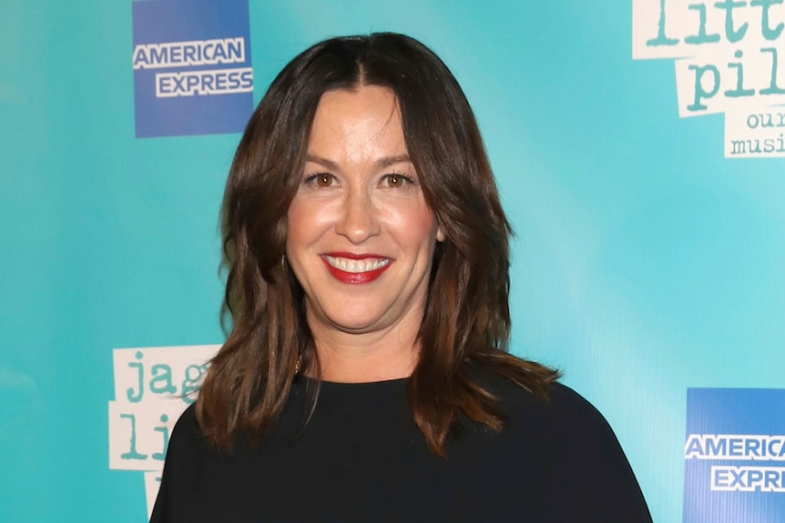 Alanis Morissette stands in front of a press sign which says "Jagged Little Pill"