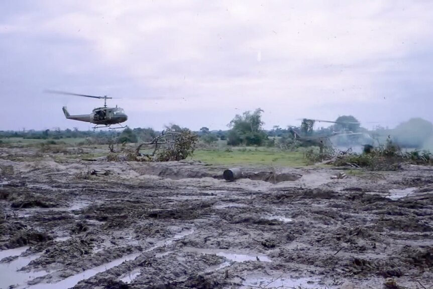Two army helicopters fly low over a muddy field.
