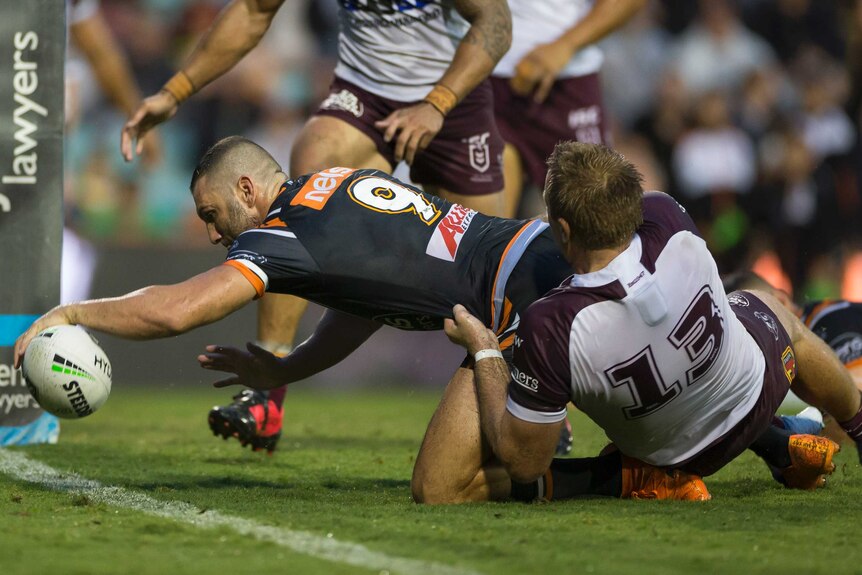 An NRL player reaches out to plant the ball down for a try as an opponent tackles him near the line.
