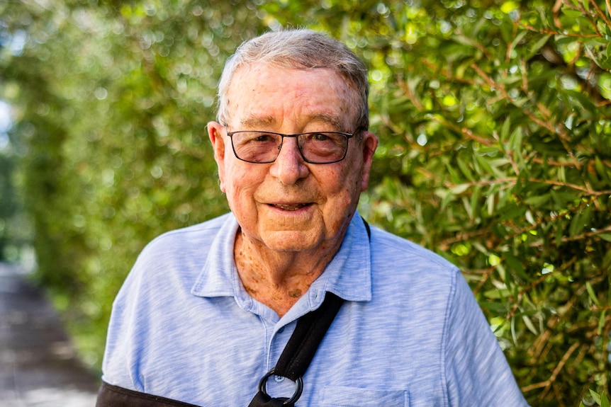 An elderly man wearing an arm brace, blue shirt and reading glasses stands beside a hedge, smiling.