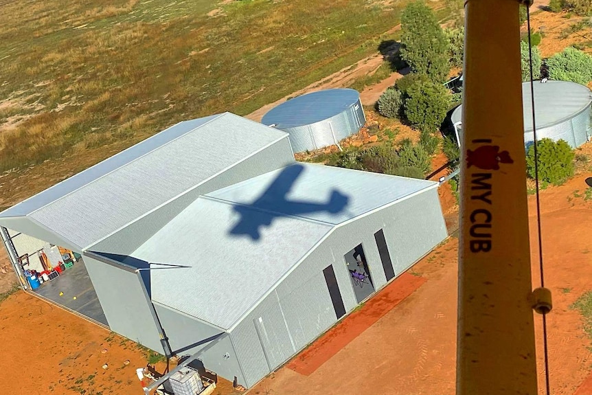Two large grey hangars on red dirt surrounded by patchy grass on a sunny day.