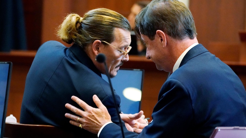 Two white middle aged men in suits huddle in a courtroom