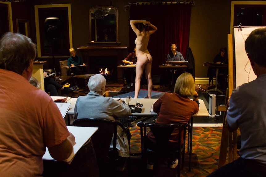 Woman standing nude in a pub room surrounded by people drawing.