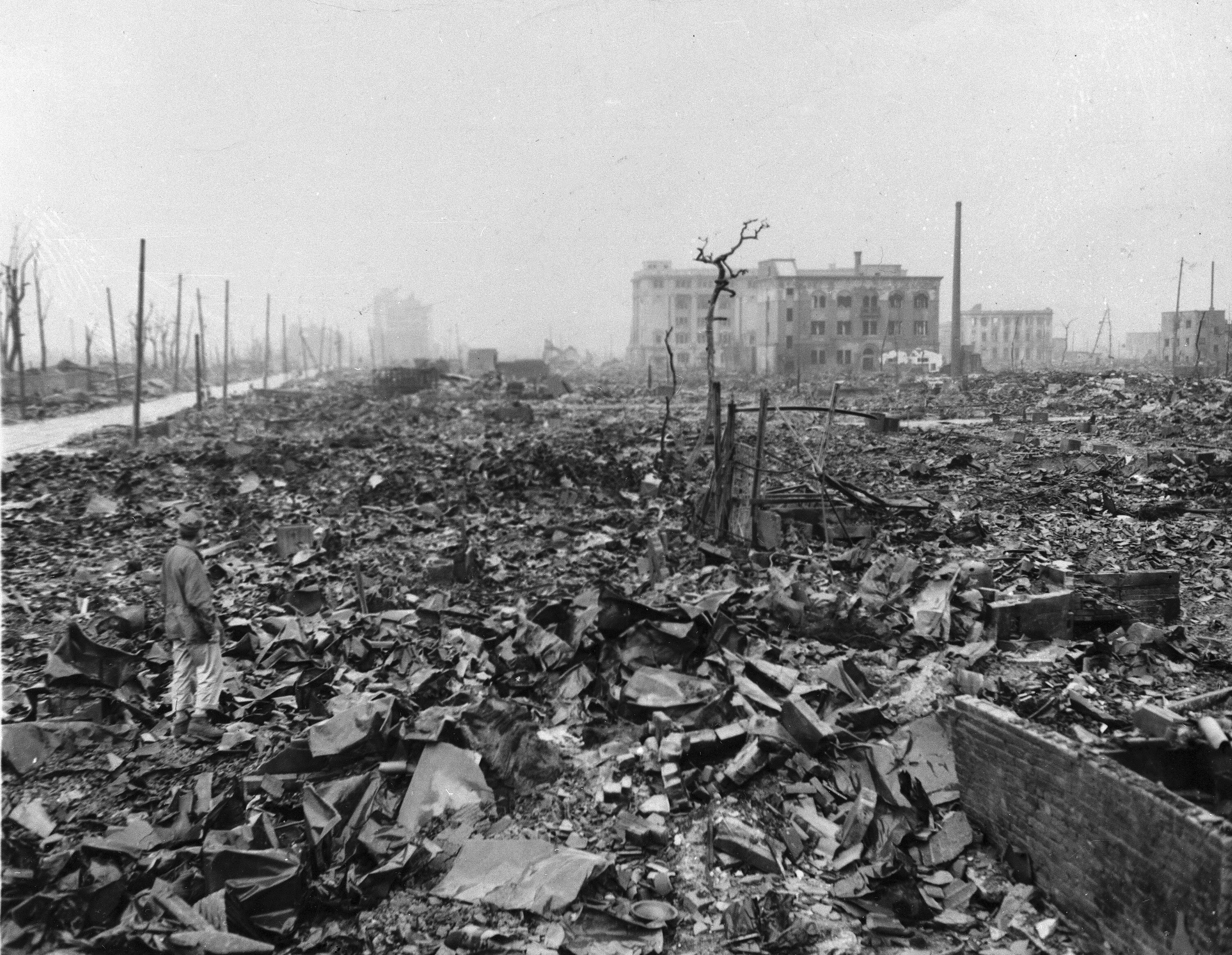 A greyscale image of a man standing on piles of rubble and destruction alongside a tree stump stripped of its leaves