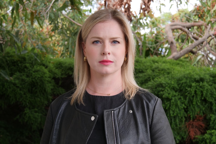 A serious blonde woman in a black leather jacket stands in front of trees and a bush.