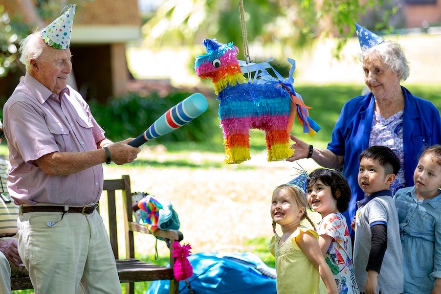 An older man (left) swings at a pinata in the park while surrounded by children.