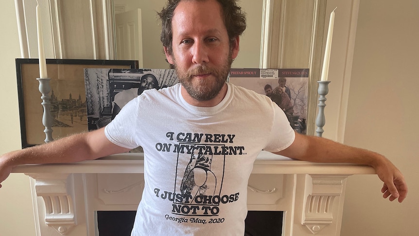 Ben Lee stands in front of fireplace, arms outstretched. He wears a shirt saying I Can Rely On My Talent I Jus Choose Not To