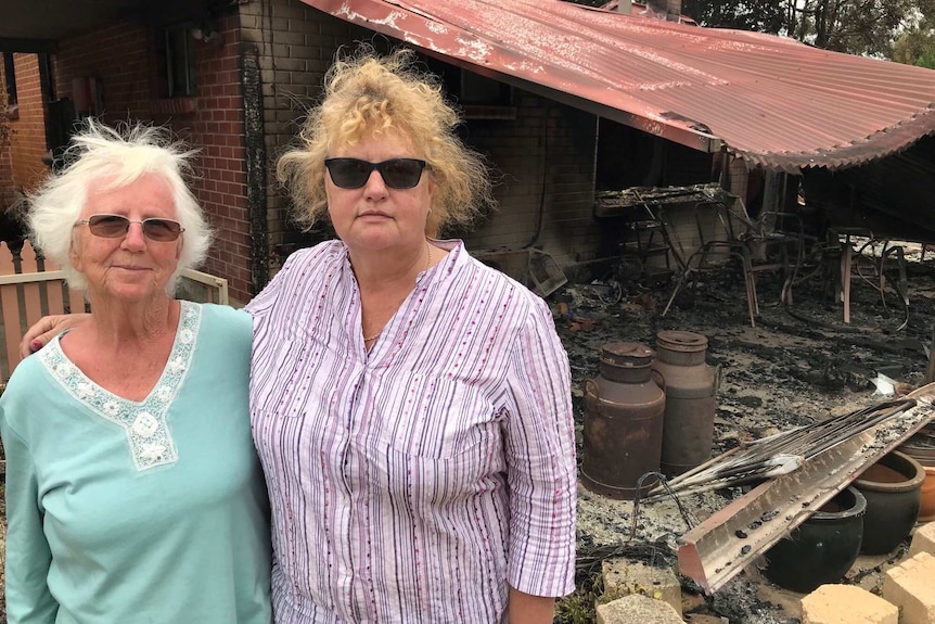 Two women wearing sunglasses stand in front of a burnt out house
