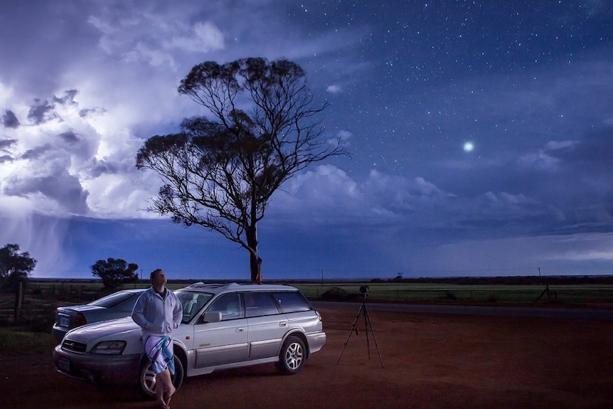 Man stands beside car on blue stormy night with tree and stars