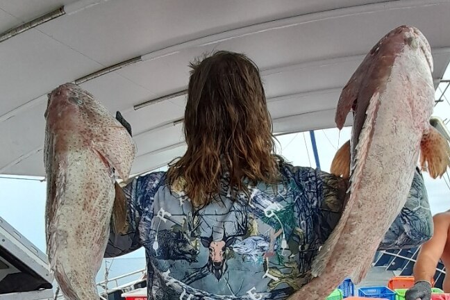 A man with a long mullet holds two large fish over his back