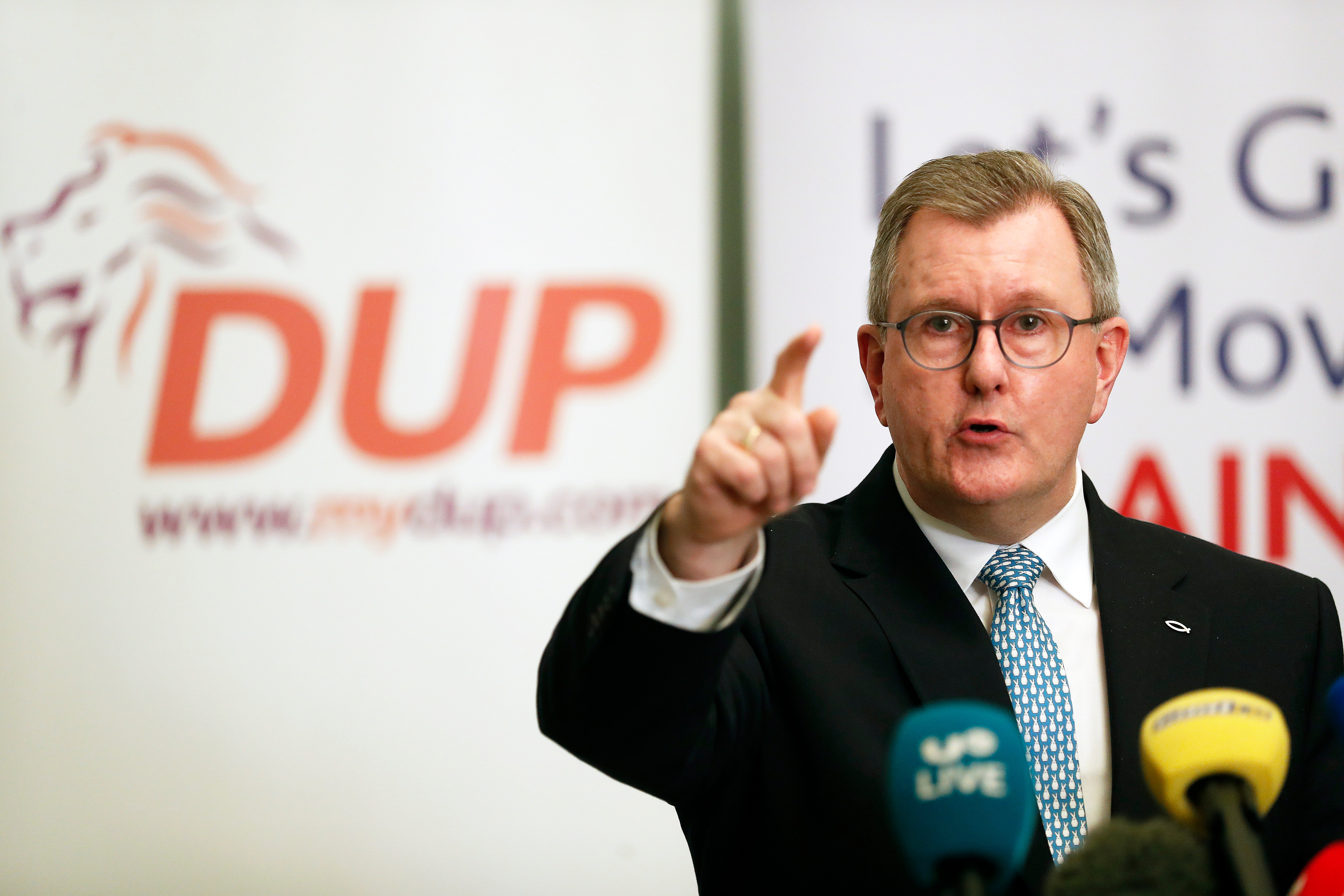 A man in a suit and tie speaks into microphones and gestures with one hand, in front of a DUP sign behind him.