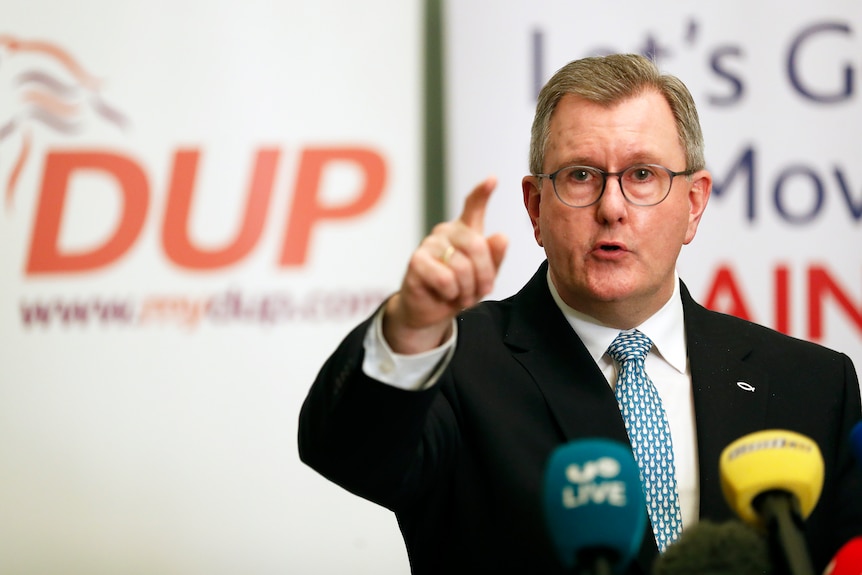 A man in a suit and tie speaks into microphones and gestures with one hand, in front of a DUP sign behind him.