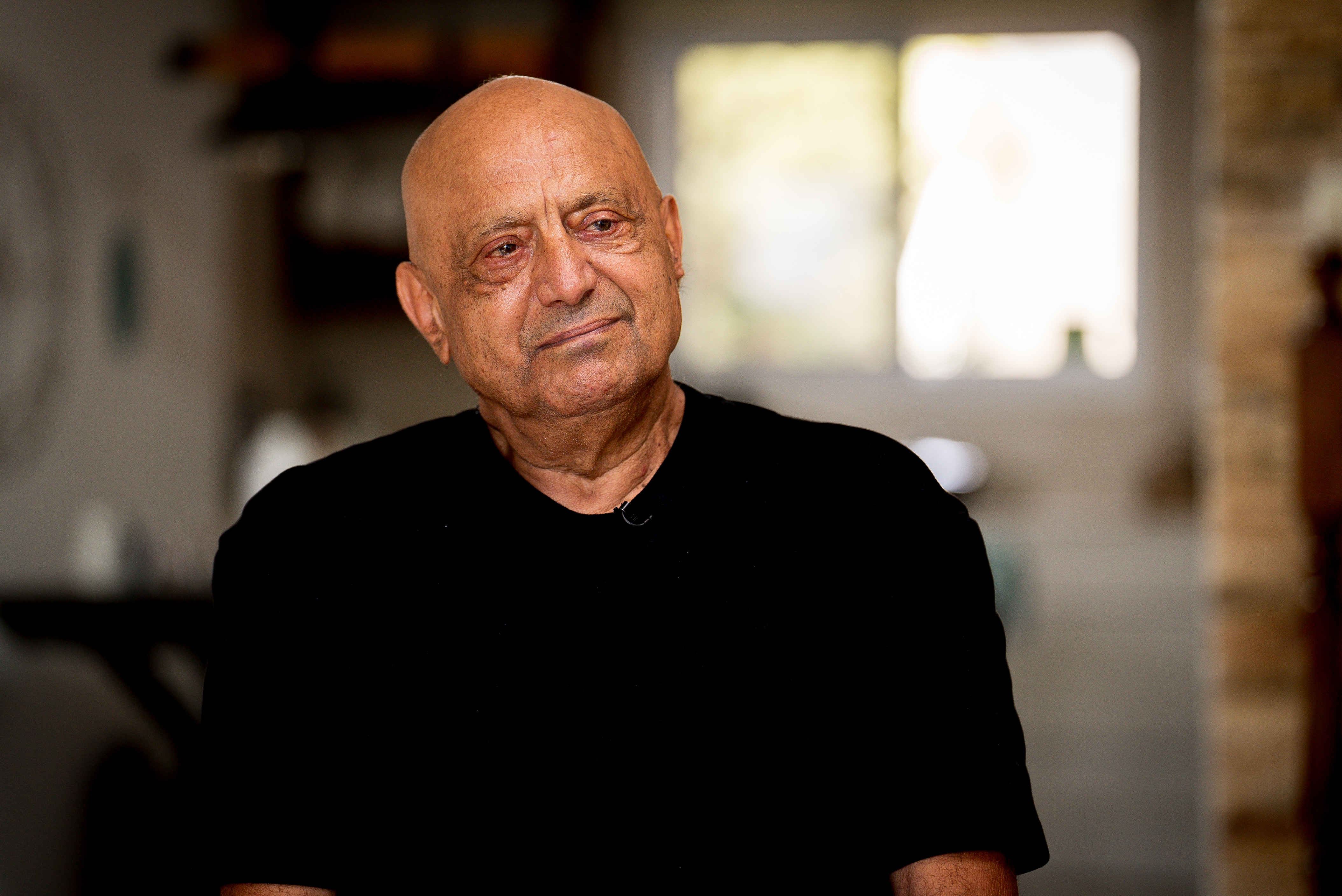 Shimon Kamari, a bald Israeli man, sits on a chair in a kitchen.
