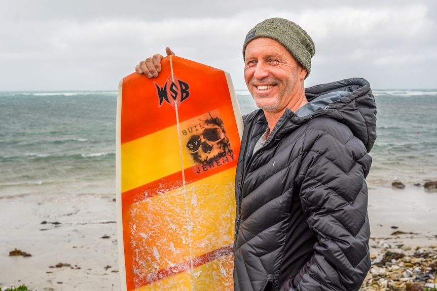 A man in a beanie and thick jacket stands by the beach holding a yellow and orange surfboard, smiling.