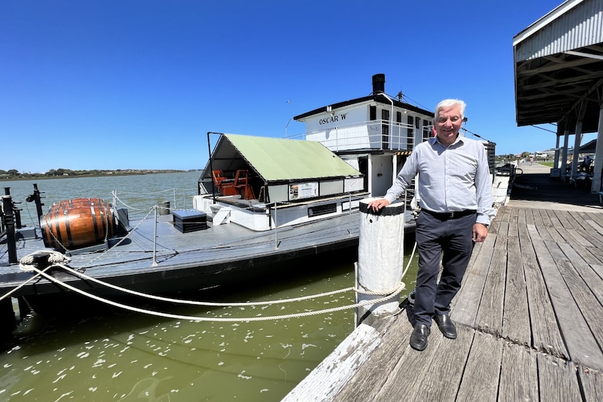 A man in a blue shirt and navy slacks stands on a wooden wharf in front of a paddle steamer boat