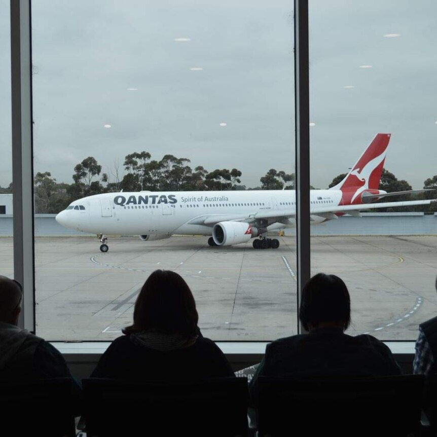 A Qantas plane taxies along a runway in overcast conditions while passengers seated inside watch on.