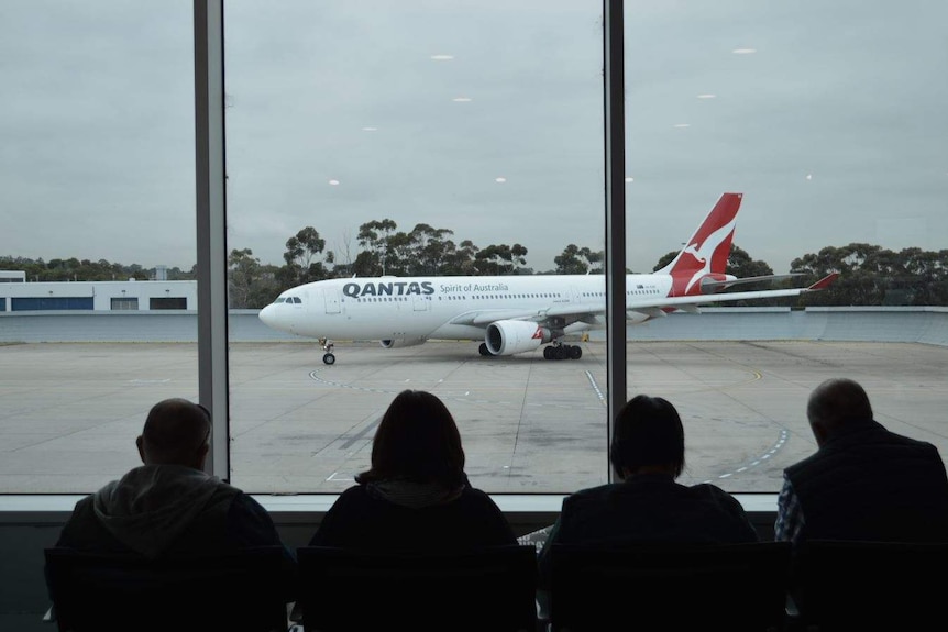 A Qantas plane taxies along a runway in overcast conditions while passengers seated inside watch on.